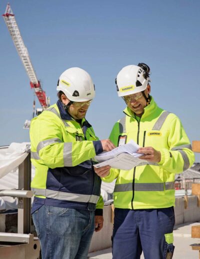 Kaksi työvaatteisiin pukeutunutta miestä katsoo yhdessä paperia työmaalla.