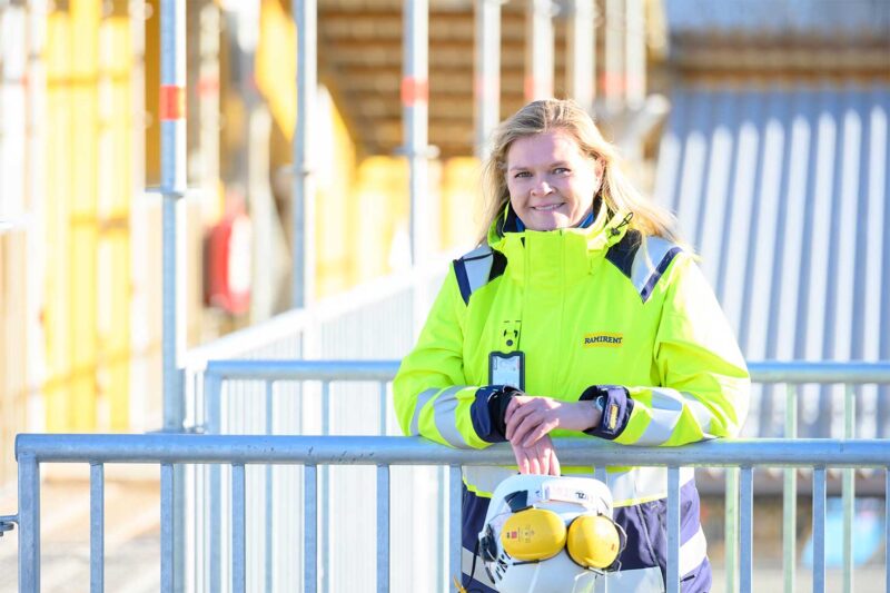 Työvaatteisiin pukeutunut nainen nojaa aitaan työmaalla ja katsoo kameraan.