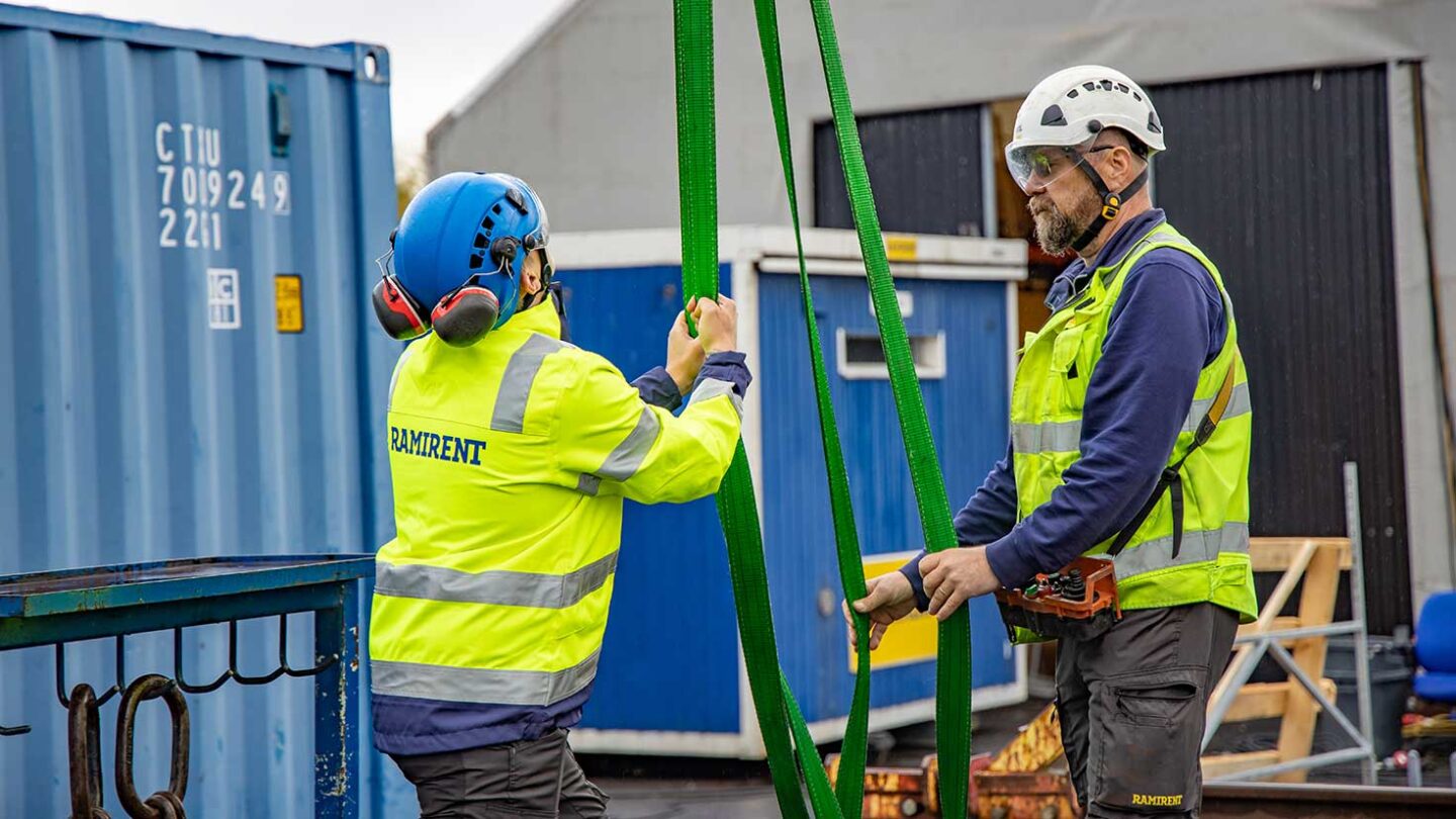 Kaksi kypäräpäistä miestä työtakeissa kiinnittää liinoja nostettavaan kohteeseen.
