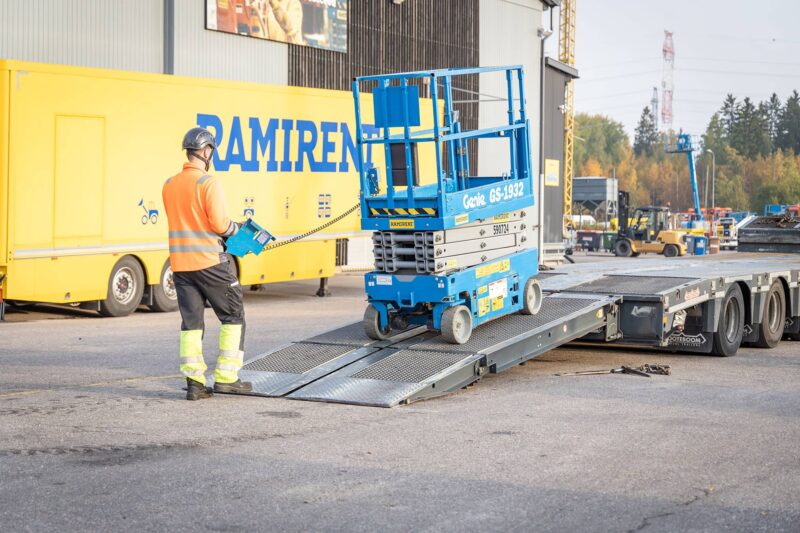 Kypäräpäinen mies lastaa sinistä saksinostinta perävaunuun kulkemalla itse takana ja ohjaamalla nostinta ohjausyksiköllä.