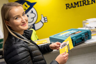 Pitkähiuksinen nainen seisoo asiakaspalvelutiskillä ja katsoo hymyillen kameraan.