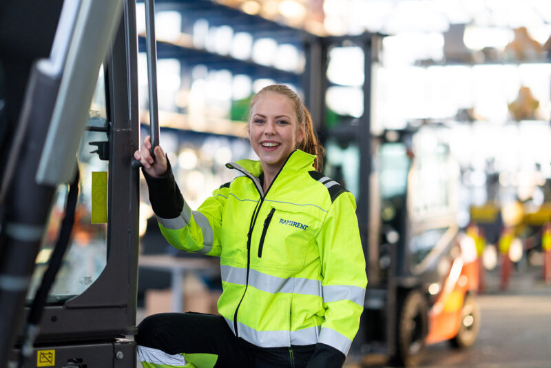 Työvaatepukuinen hymyilevä nainen pitää kiinni trukin kahvasta ja katsoo hymyillen kameraan.