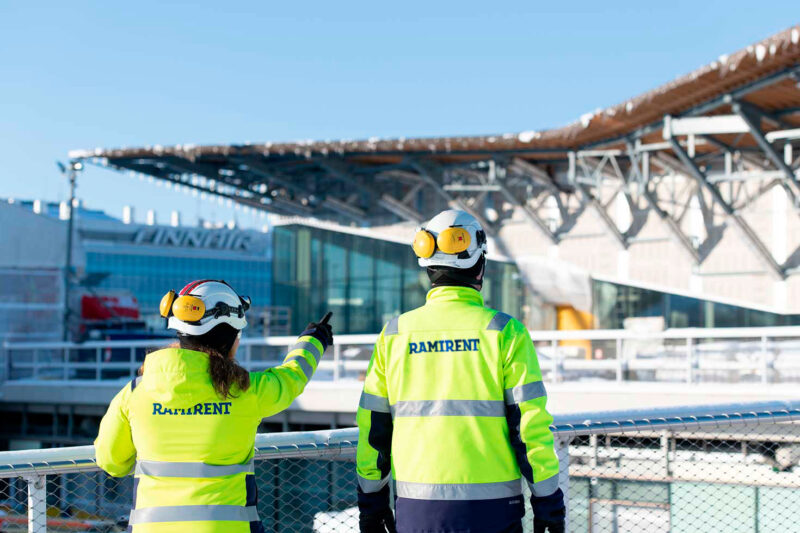 Kaksi työvaatepukuista ihmistä seisoo selin kameraan ja katsoo taustalla näkyvää terminaalityömaata.