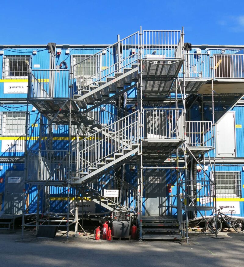 Sinisiä työmaatiloja kolmessa kerroksessa ja niiden edessä oleva porrastorni.