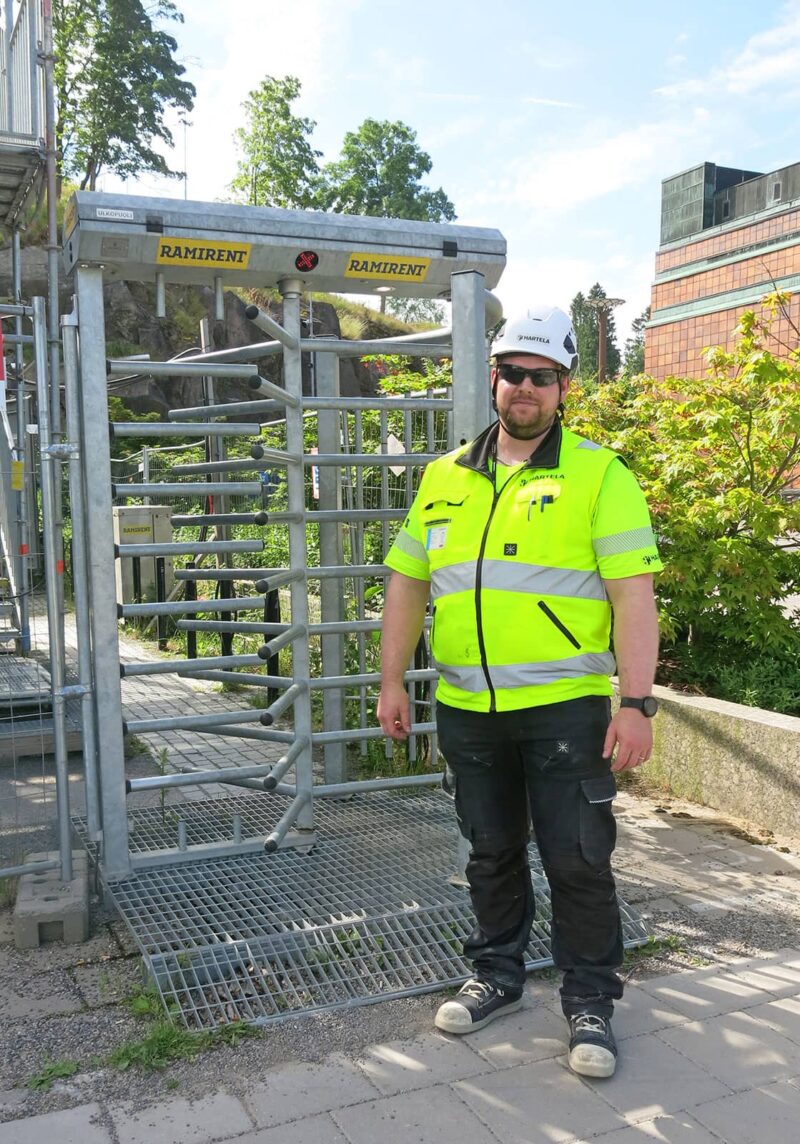 Työvaatteisiin ja kypärään pukeutunut mies seisoo työmaan kulkuportin edessä.