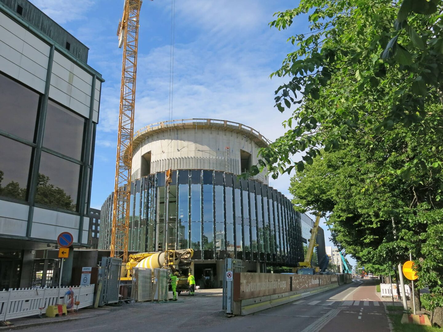 Kaupunkikuvassa näkyy työmaa aitoineen sekä rakenteilla oleva pyöreämutoinen korkea rakennus.