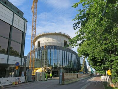 Kaupunkikuvassa näkyy työmaa aitoineen sekä rakenteilla oleva pyöreämutoinen korkea rakennus.