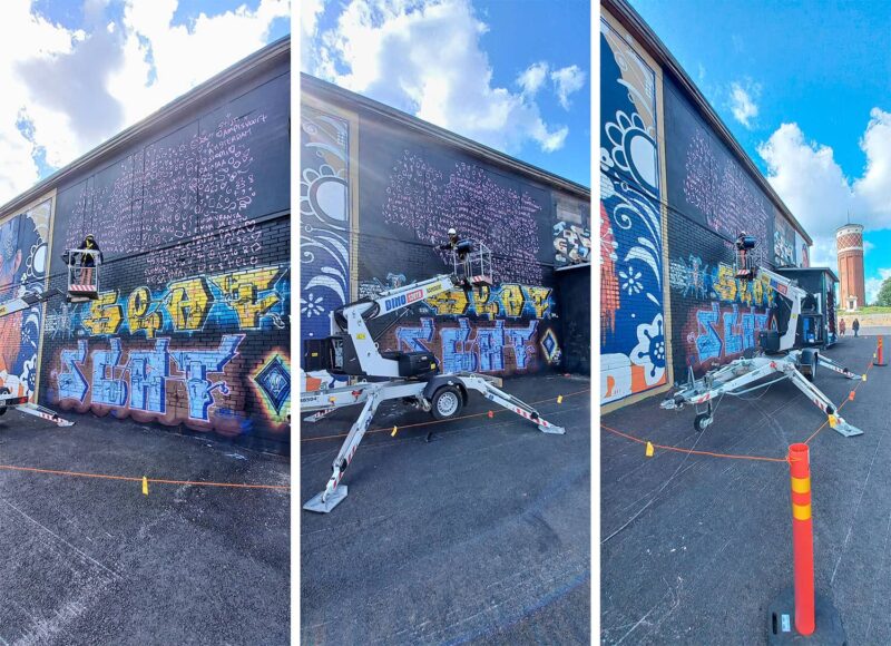 Kolme vierekkäistä kuvaa naisesta henkilönostimen korissa maalaamassa suurta kuvaa rakennuksen seinään.