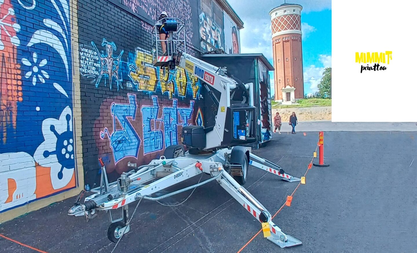 Nainen seisoo henkilönostimen korissa ja maalaa suurta kuvaa rakennuksen seinään.