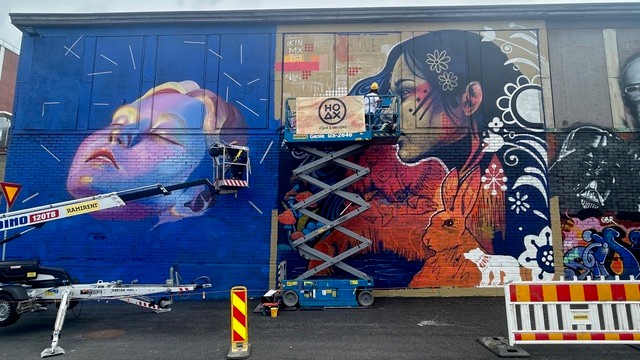 Teleskooppipuominostin ja saksinostin ison maalattavana olevan seinän edessä. Tekeillä on erilaisia muraalimaalauksia.