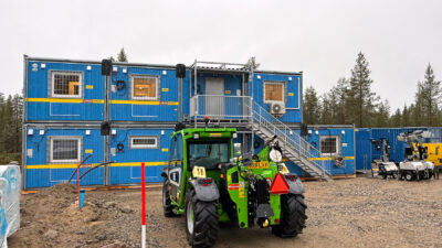 Vihreä kurottaja seisoo työmaalla kahteen kerrokseen asennettujen sinisten työmaatilojen edessä.