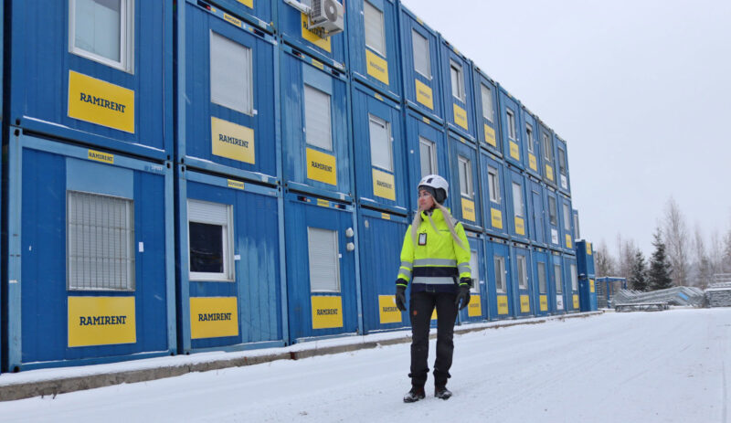 Työvaatteisiin pukeutunut nainen seisoo kolmikerroksisen sinisen työmaatilarivistön edessä lumisella pihalla.