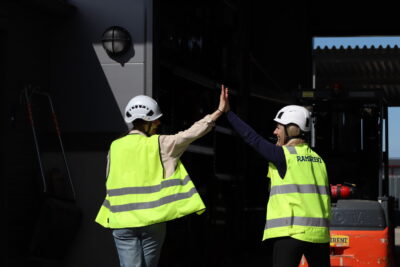 Kaksi huomioliiveihin ja kypäröihin sonnustautunutta nauravaa naista lyö kädet yhteen takaapäin kuvattuna.