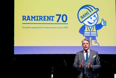 Harmaaseen pukuun sonnustautunut mies puhuu lavalla ja taustalla näkyy Rami-hahmo ja teksti Ramirent 70.