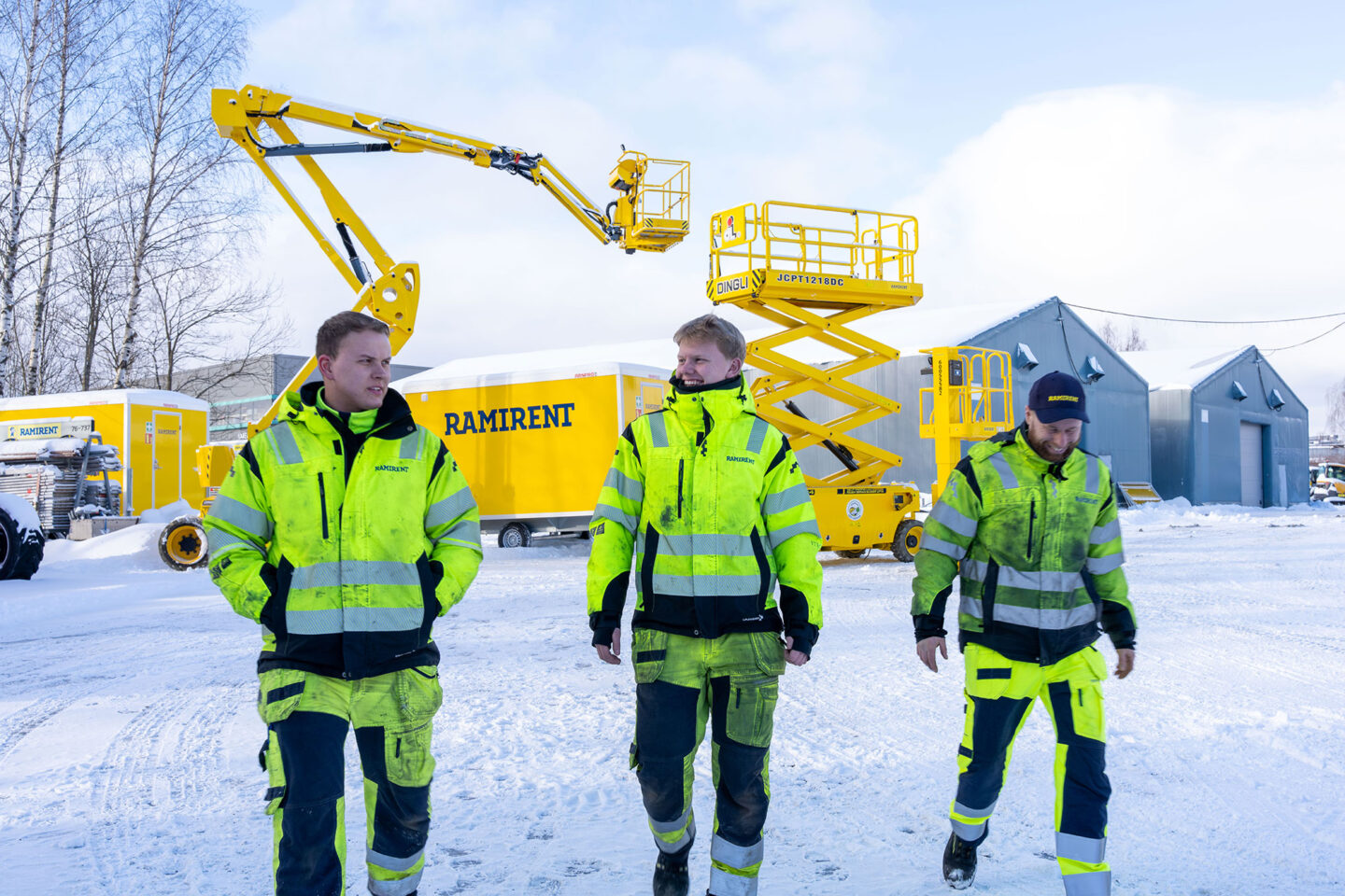 Kolme työvaatepukuista miestä kävelee lumisella konevuokraamon pihalla taustalla keltaisia henkilönostimia.