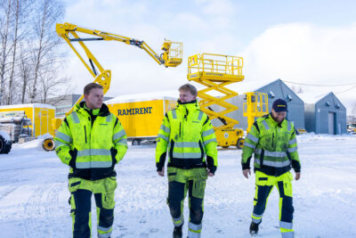 Kolme työvaatepukuista miestä kävelee lumisella konevuokraamon pihalla taustalla keltaisia henkilönostimia.