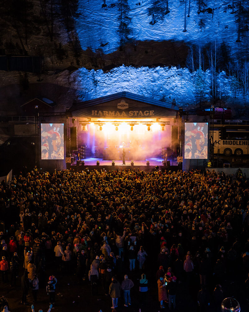 Tummasävyinen talvinen kuva esiintymislavasta jonka edessä on suuri ihmisjoukko ja takana valaistuja puita ja laskettelurinne.