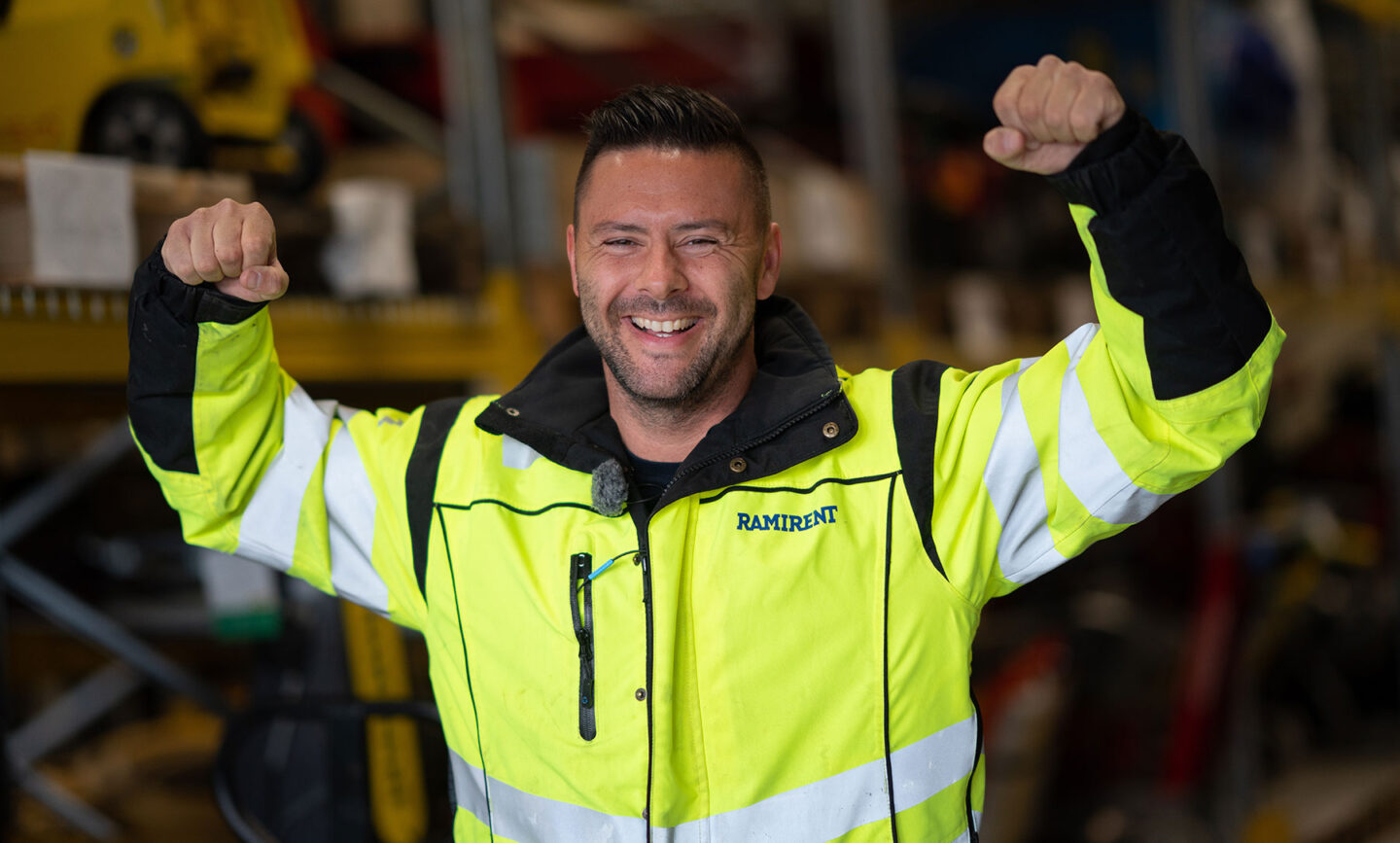 Iloinen mies tuulettaa käsillään konevuokraamon varastohyllyjen välissä.