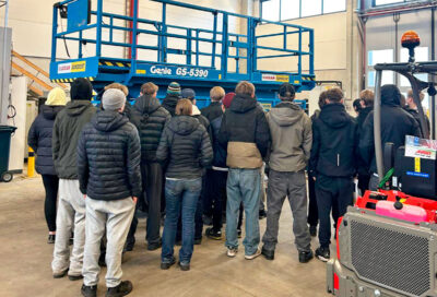 Joukko nuoria opiskelijoita seisoo saksinostimen ympärillä konevuokraamon hallissa.