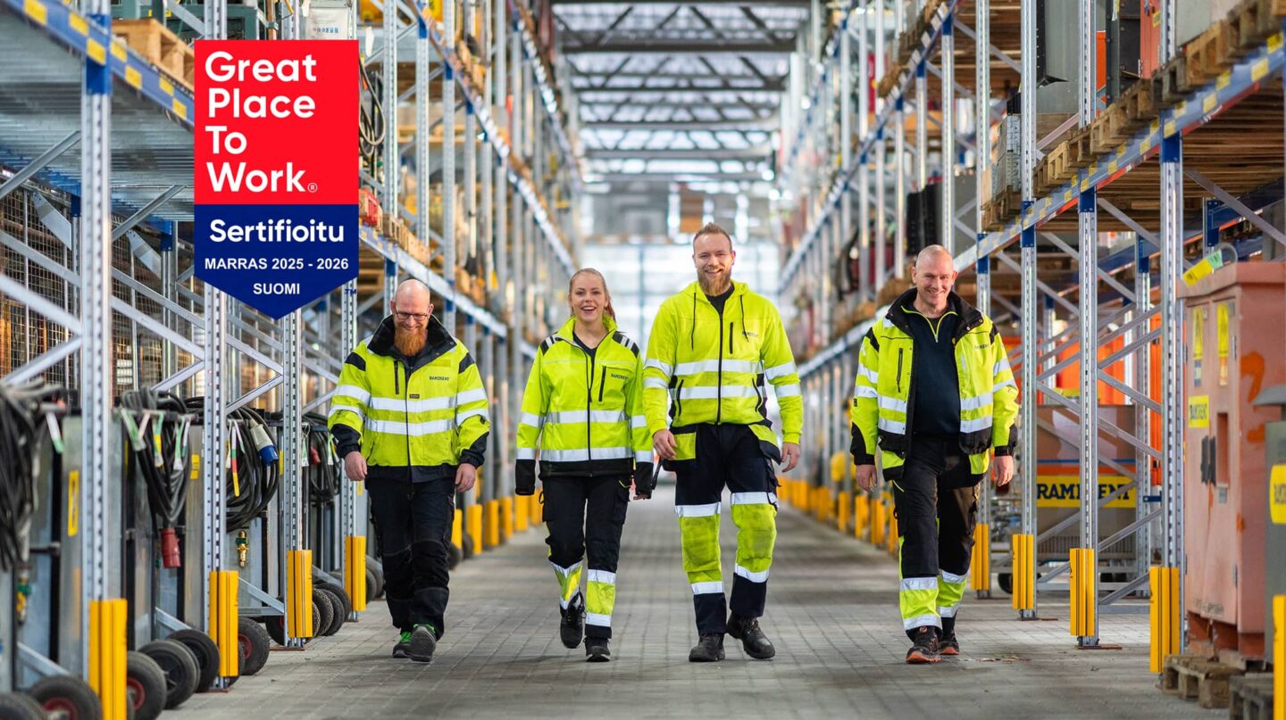 Neljä työvaatepukuista hymyilevää henkilöä kävelee kohti kameraa konevuokraamon varastokäytävällä. Kuvassa on myös Great Place to Work sertifikaattilogo.