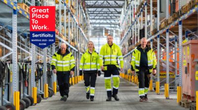 Neljä työvaatepukuista hymyilevää henkilöä kävelee kohti kameraa konevuokraamon varastokäytävällä. Kuvassa on myös Great Place to Work sertifikaattilogo.