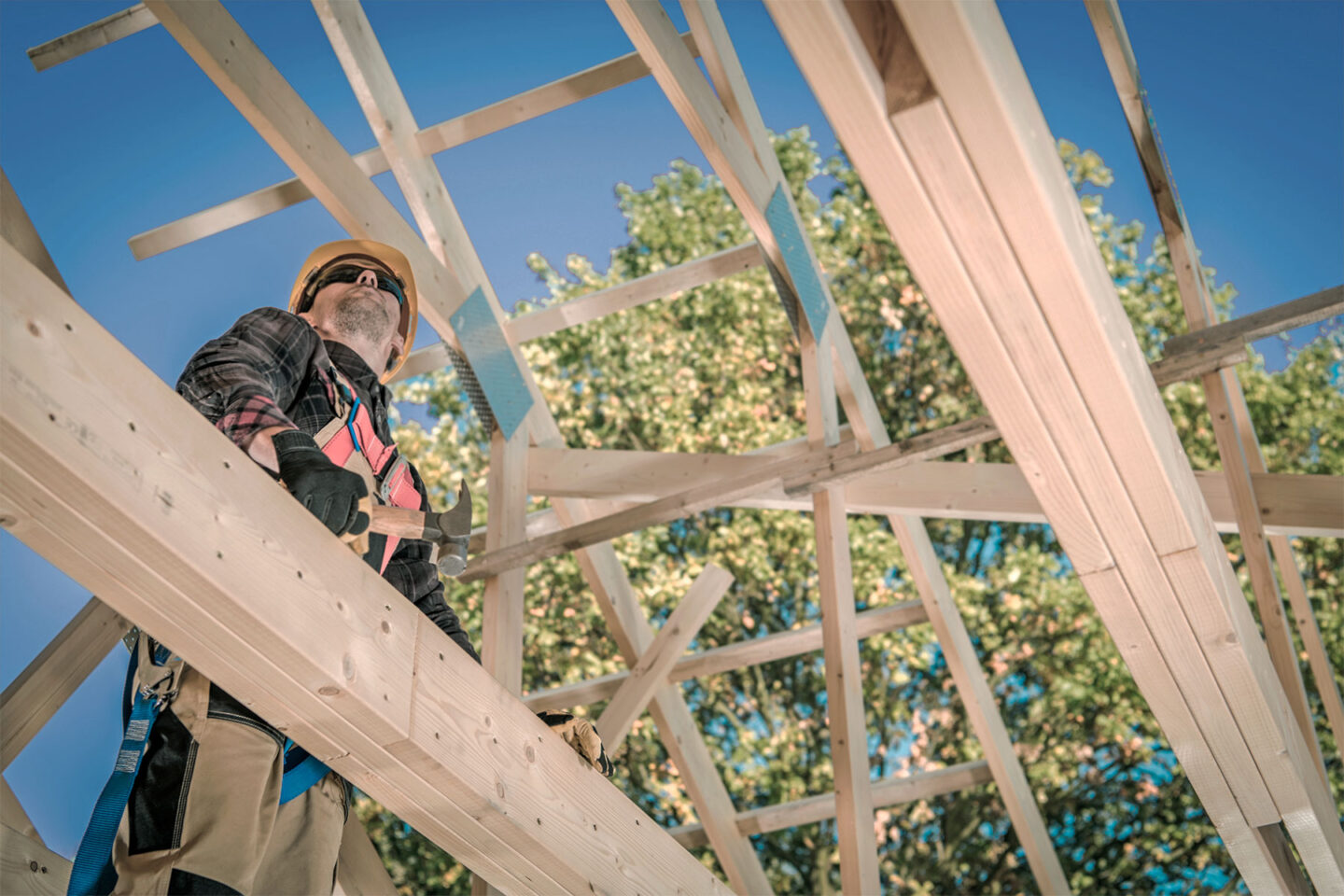 Kypäräpäinen mies seisoo vasara kädessä talon puurakenteissa alhaaltapäin kuvattuna.