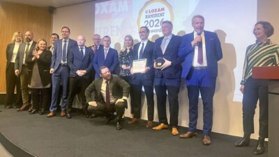 Rivi miehiä ja naisia lavalla valkokankaan edessä seisomassa ja yksi miehistä puhuu mikrofoniin.