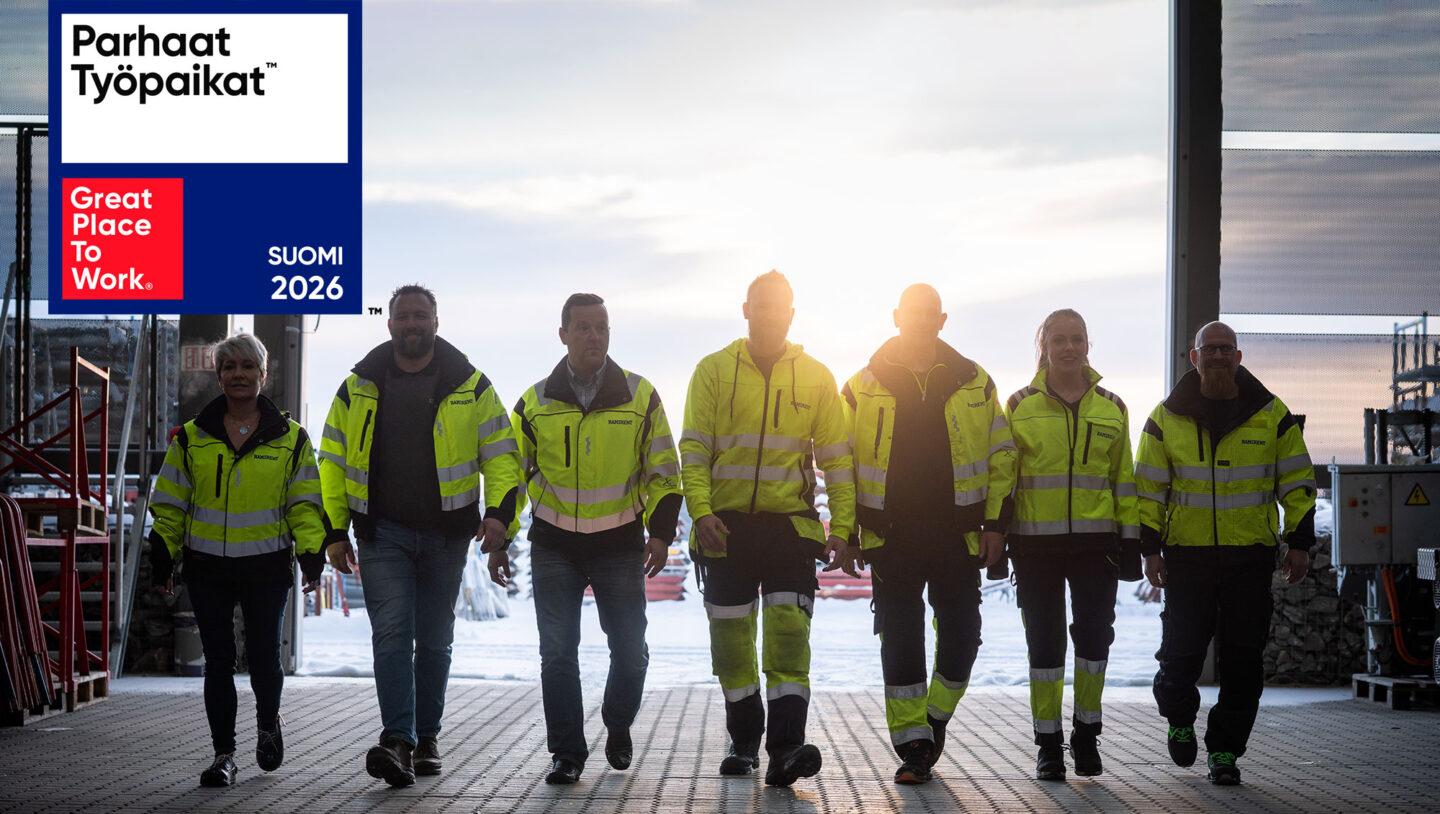 Joukko työvaatteisiin pukeutuneita ihmisiä kävelee kohti kameraa varastohallissa.