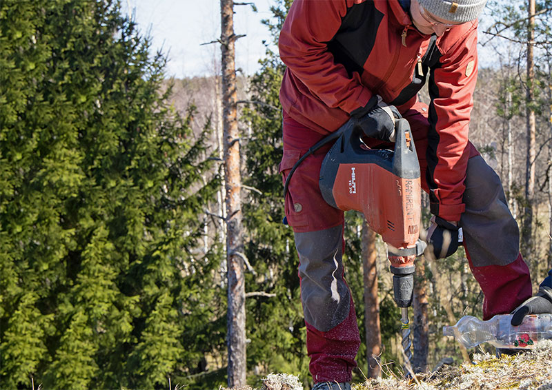 Punaisiin pukeutunut mies poraa kalliota metsäisessä maisemassa.