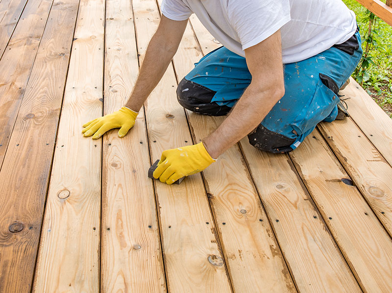 Työhousuihin ja t-paitaan pukeutunut henkilö on polvillaan terassilautojen päällä.